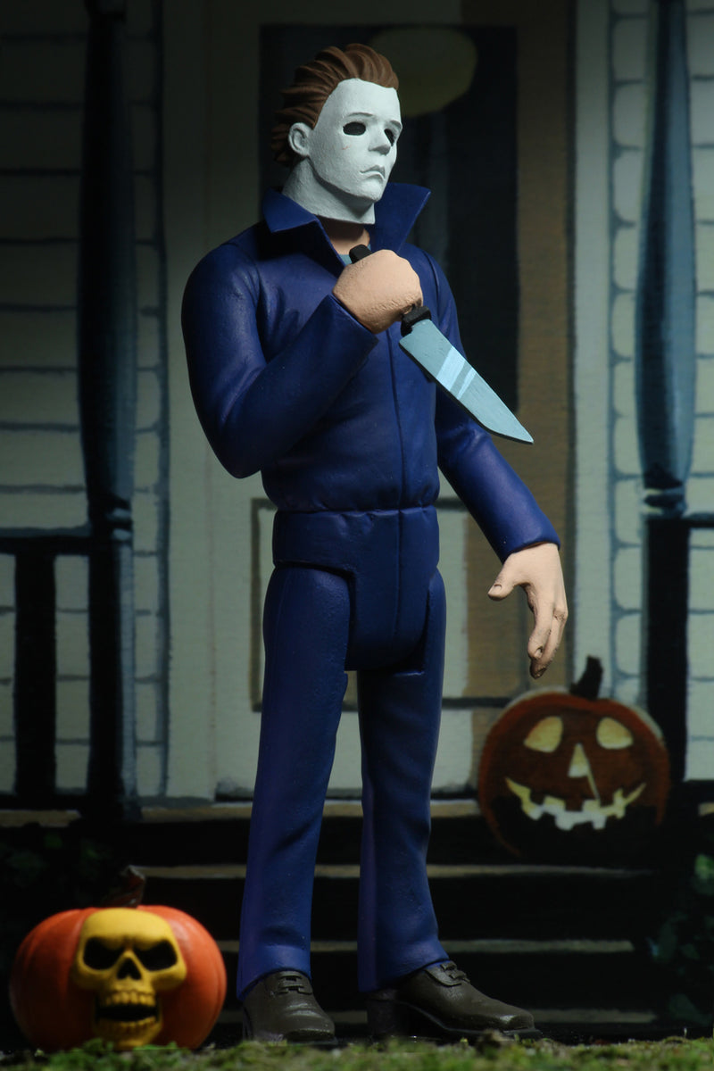 Michael Myers action figure is standing in blue coveralls on a front porch of a house in Haddonfield, holding a knife, with a pumpkin at his feet.