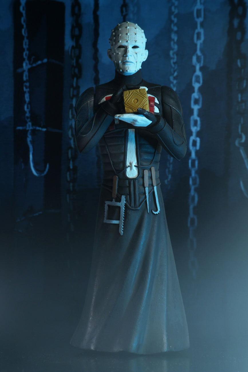 Pinhead is standing in a black dress outfit with tools hanging from it, holding a brown box, with blue lighting.