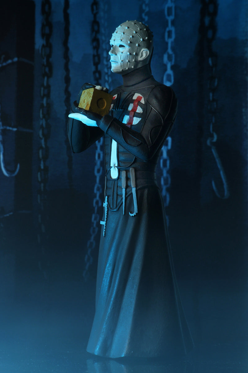 Pinhead action figure is standing in a black dress outfit with tools hanging from it, holding a brown box, with blue lighting.