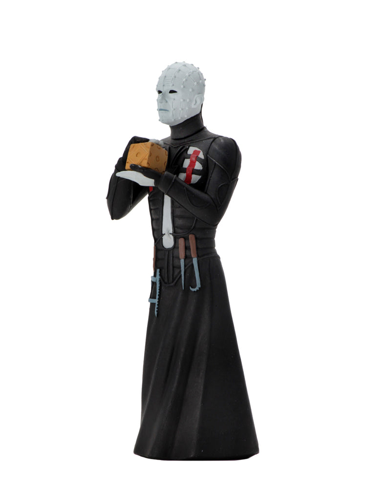 Pinhead NECA action figure is standing in a black dress outfit with tools hanging from it, holding a brown box, with white lighting.