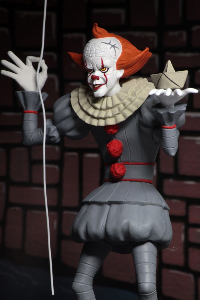 This is a NECA Toony Terrors It 2017 Pennywise action figure and he is wearing a grey clown suit that has three red balls and a white collar, with white gloves, balls on his shoes, a white face and orange hair and is holding a white boat and balloon string.