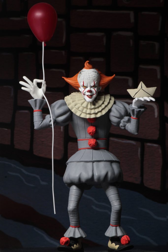 This is a NECA Toony Terrors It 2017 Pennywise action figure and he is wearing a grey clown suit that has three red balls and a white collar, with white gloves, balls on his shoes, a white face and orange hair and is holding a white boat and red balloon.