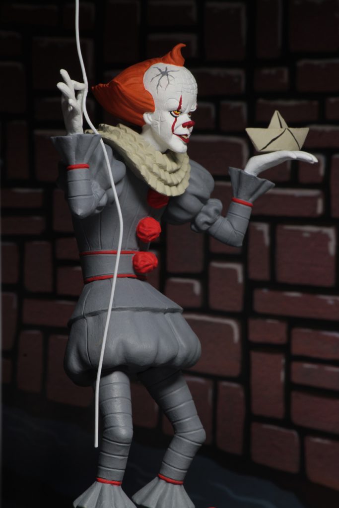 This is a NECA Toony Terrors It 2017 Pennywise action figure and he is wearing a grey clown suit that has three red balls and a white collar, with white gloves, balls on his shoes, a white face and orange hair and is holding a white boat.
