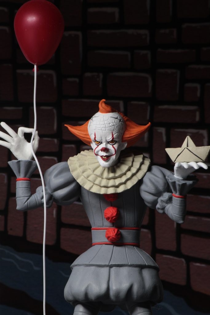 This is a Toony Terrors It 2017 movie of a Pennywise the clown posable NECA 6" action figure, who is wearing a grey clown suit with red balls, white gloves, holding a red ballon and who has a red nose, white face and orange hair.