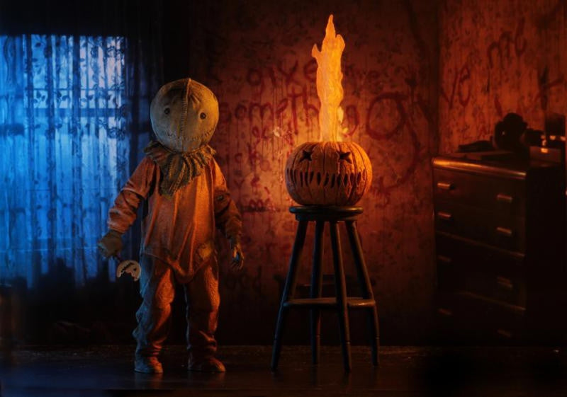 This is a Trick R Treat Sam NECA 7" ultimate action figure and there is a on orange pumpkin on a stool with the teeth and eyes cut out and fire coming out of it and he is wearing an orange suit, burlap mask, gloves and is holding a bitten lollipop.