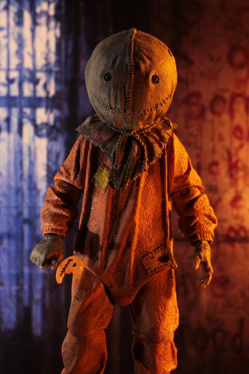 This is a Trick R Treat Sam NECA 7" ultimate action figure and he has a burlap mask, an orange suit, gloves and he is holding a bitten pumpkin lollipop.
