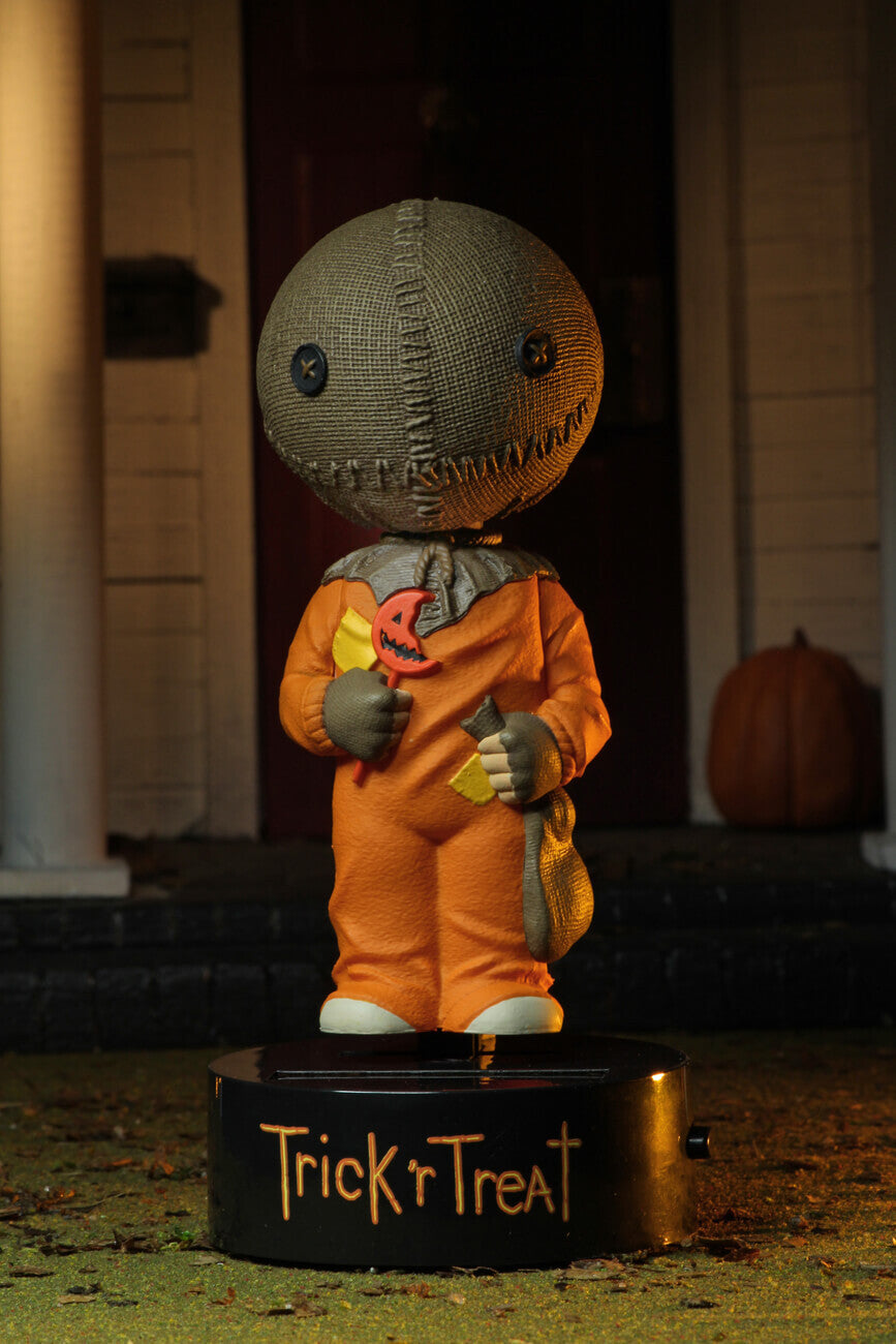This is a Trick "r Treat Sam NECA Body Knocker and he has a plastic black base, brown burlap head, orange suit and a bitten orange lollipop and pumpkin in the background.