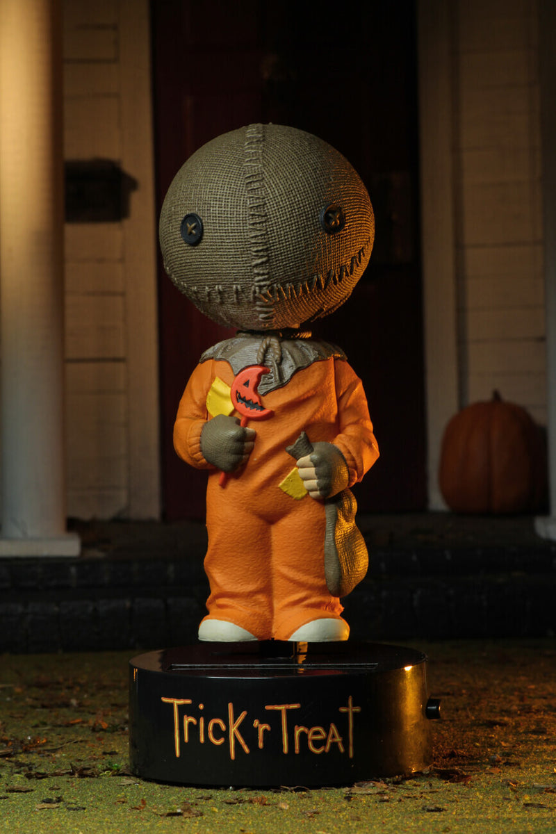 This is a Trick "r Treat Sam NECA Body Knocker and he has a plastic black base, brown burlap head, orange suit and a bitten orange lollipop and pumpkin in the background.