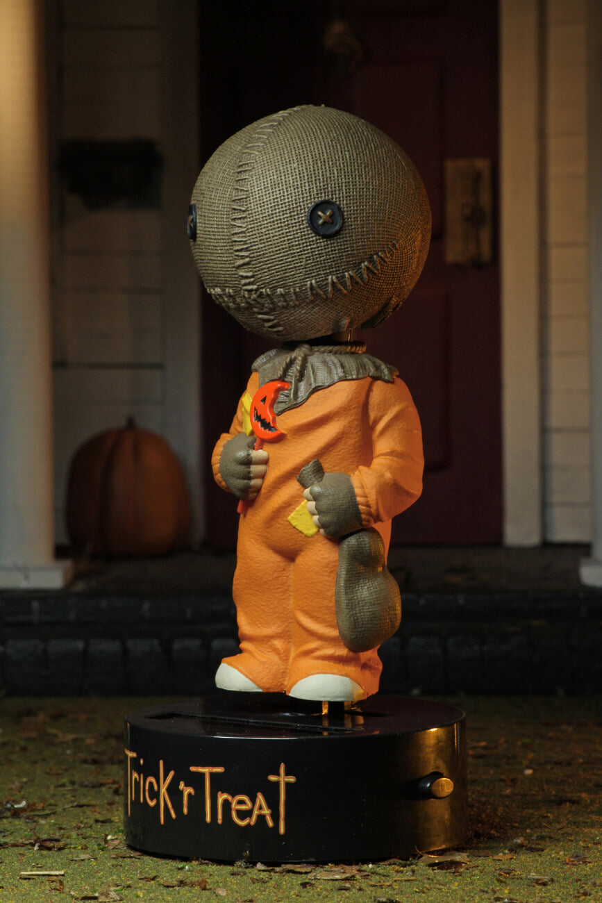 This is a Trick "r Treat Sam NECA Body Knocker and he has a plastic black base, brown burlap head, orange suit and a bitten orange lollipop and a rope on his neck.