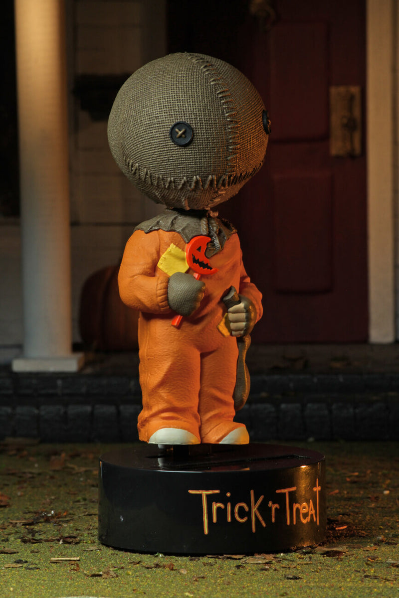 This is a Trick "r Treat Sam NECA Body Knocker and he has a plastic black base, brown burlap head, orange suit and a bitten orange lollipop in front of an yellow patch..