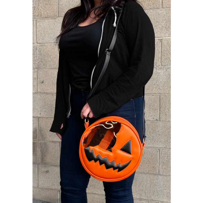 This is a Trick 'R Treat Sam bitten lollipop purse and it is orange, with a black eye and smile, has a black strap and is on a person who is wearing jeans and a black sweatshirt..