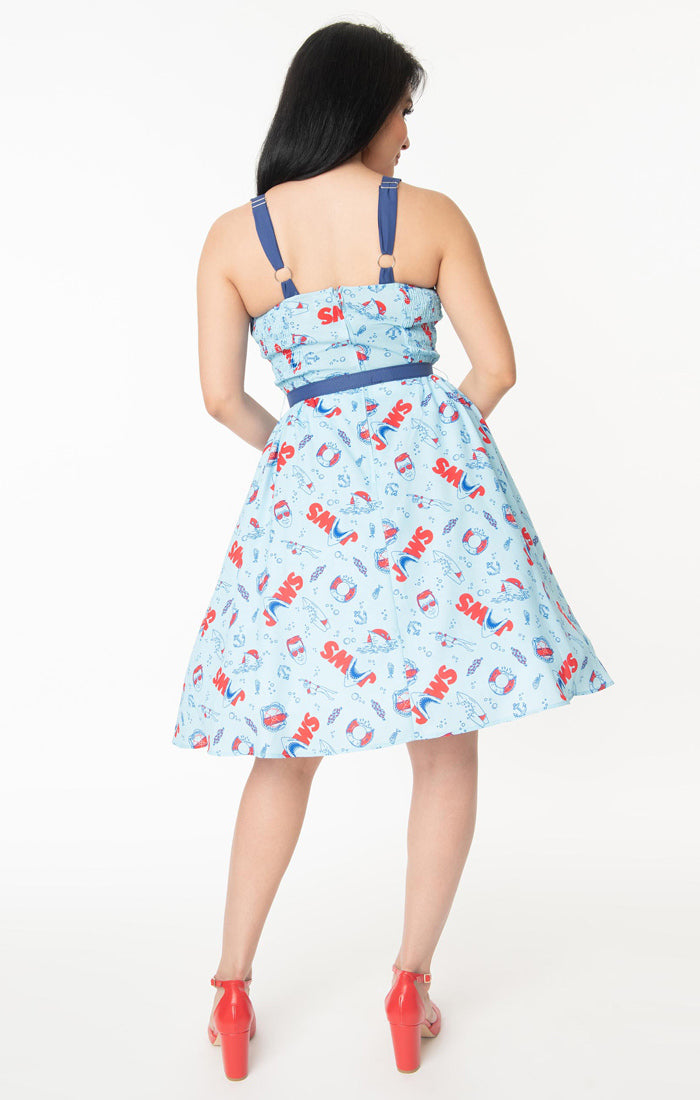 This is a Unique Vintage Jaws dress and the model has dark hair, red shoes and the dress is blue with red text and sharks on it, a blue belt and a blue sweetheart neckline.