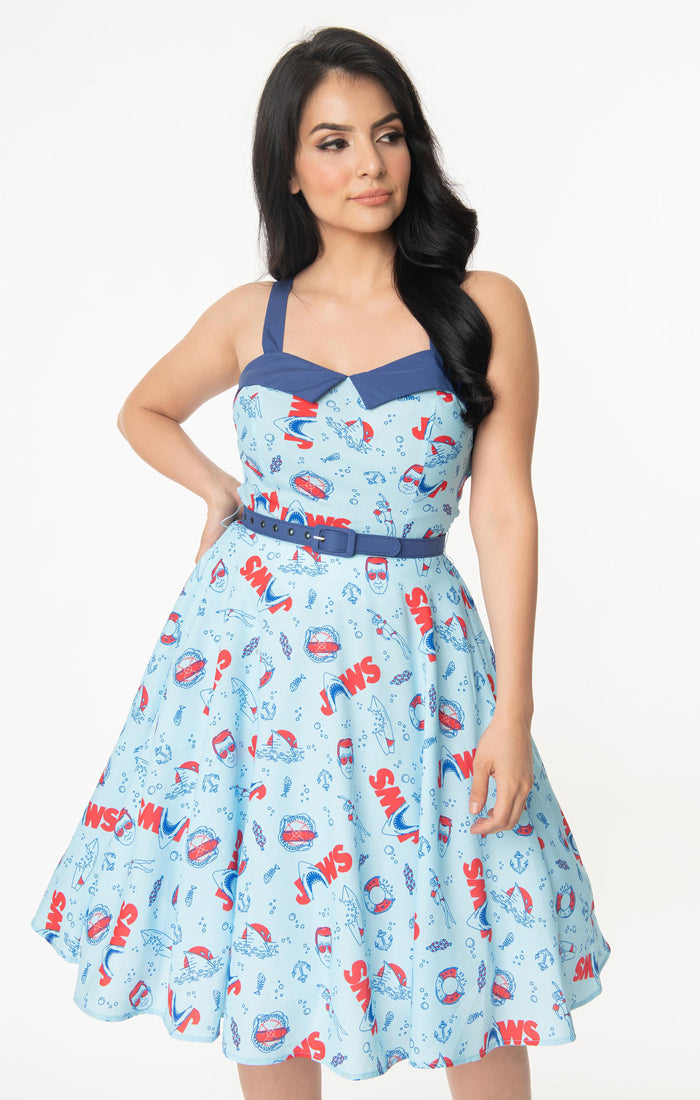 This is a Unique Vintage Jaws dress and the model has dark hair, red shoes and the dress is blue with red text and sharks on it, a blue belt and a blue sweetheart neckline.