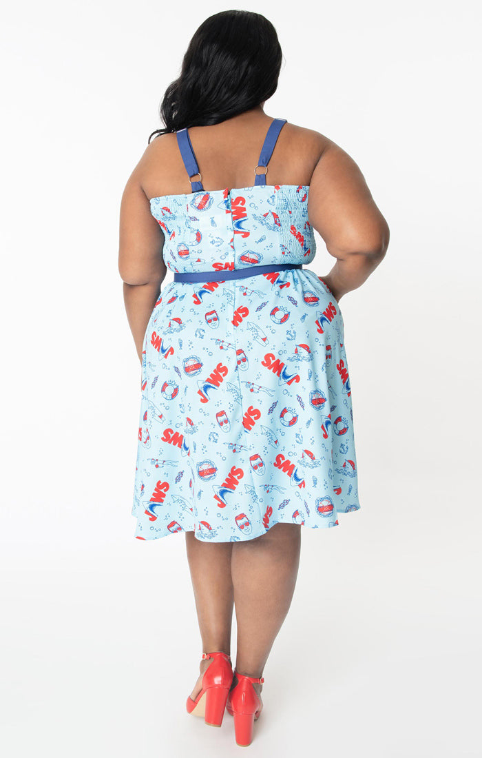 This is a Unique Vintage Jaws dress and the model has dark hair, red shoes and the dress is blue with red text and sharks on it, a blue belt and a blue sweetheart neckline on a plus size model.
