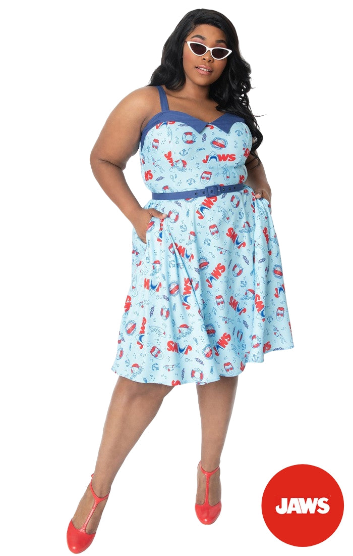 This is a Unique Vintage Jaws dress and the model has dark hair, red shoes and the dress is blue with red text and sharks on it, worn by a plus size model.
