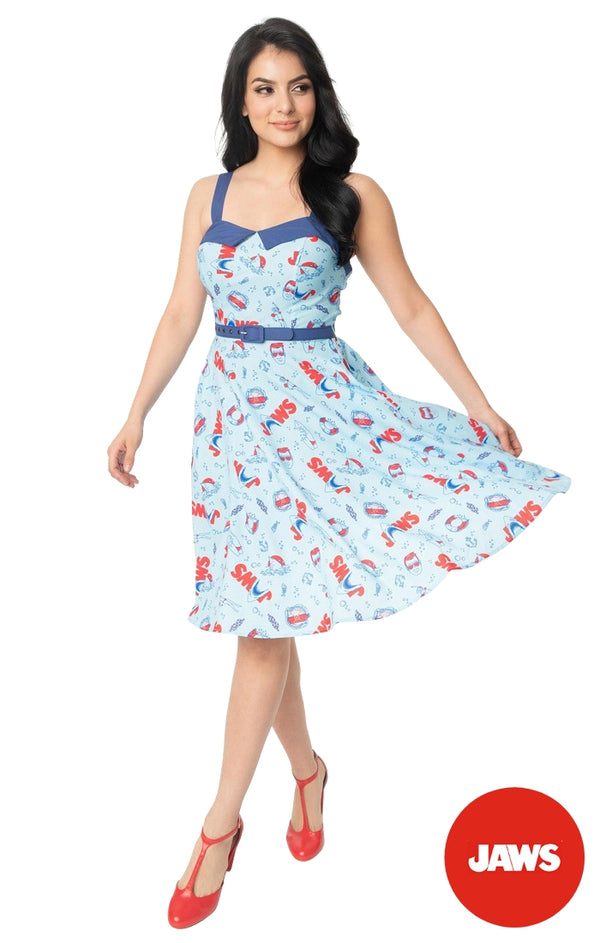 This is a Unique Vintage Jaws dress and the model has dark hair, red shoes and the dress is blue with red text and sharks on it.